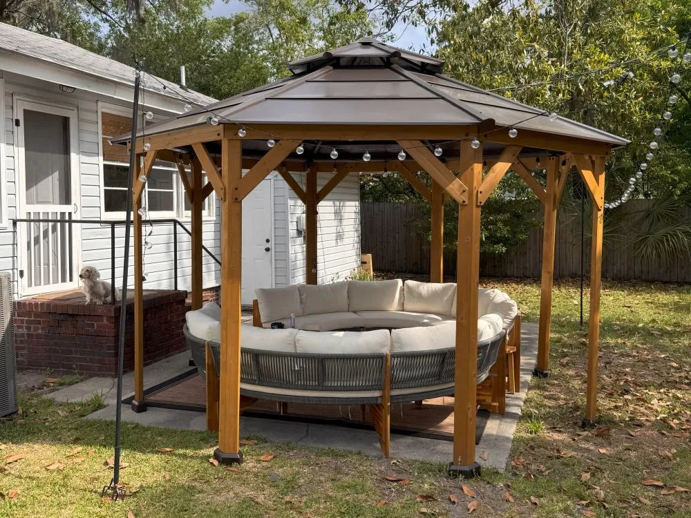 Wooden gazebo with a round sofa underneath, surrounded by a house and trees.