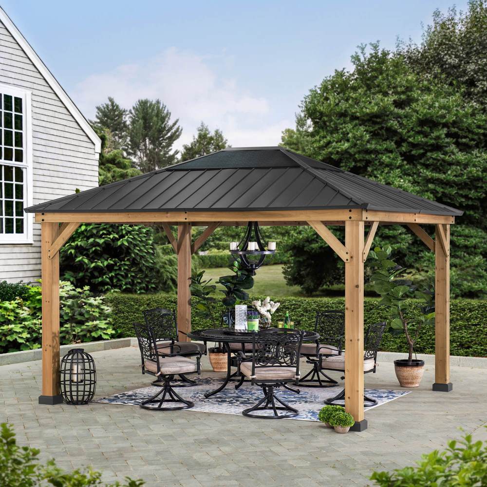 Wooden gazebo with a black metal roof on a patio, surrounded by greenery and a house.