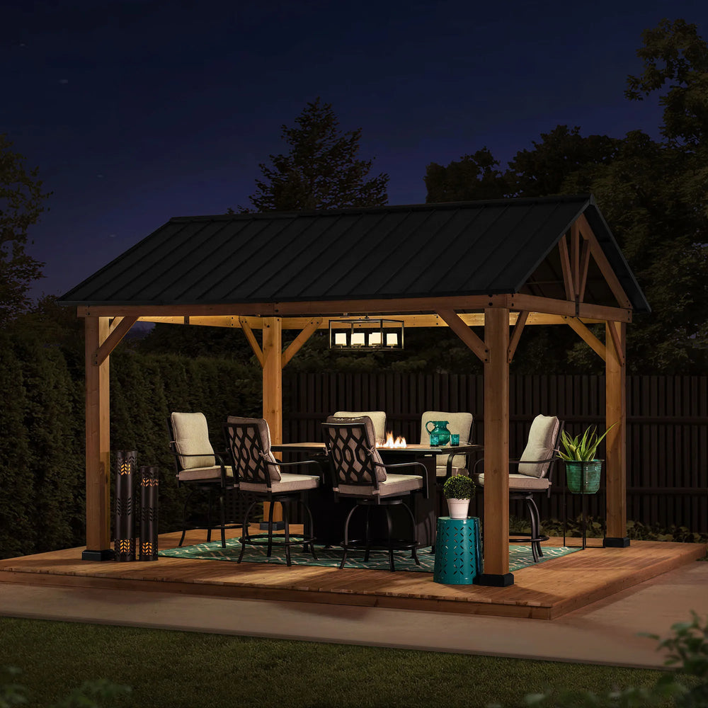 Wooden gazebo with outdoor furniture and lighting in a backyard setting at night.